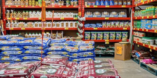Garden supplies section in a hardware store featuring stacks of potting mix and garden soil bags in the foreground. Red shelving holds fertilizers, plant food, soil conditioners, and lawn care products arranged across multiple shelves.