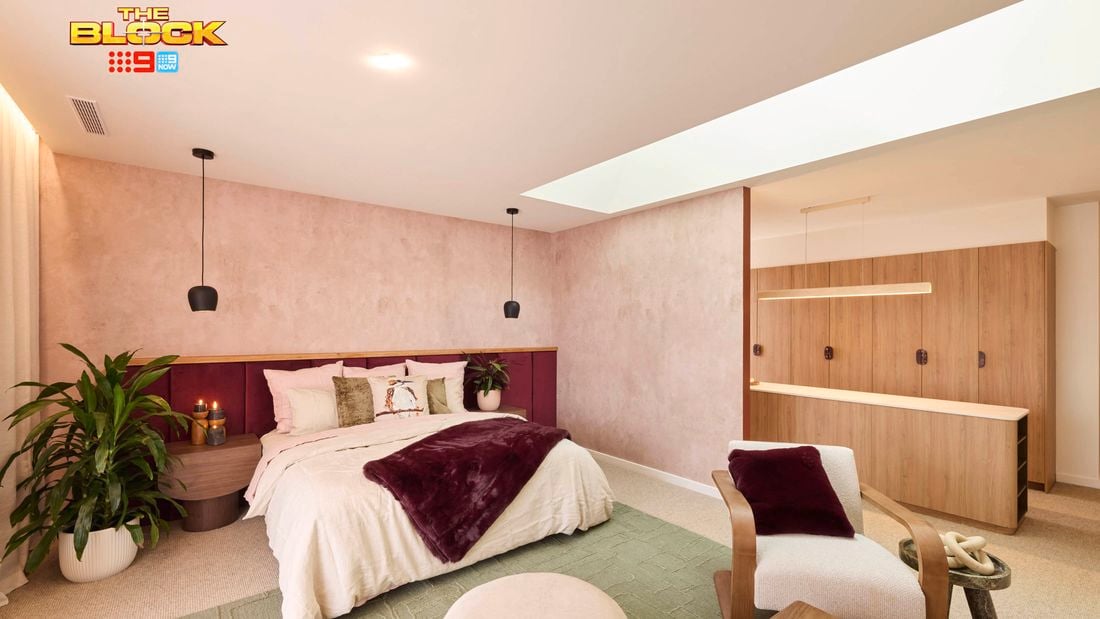 A modern bedroom with a neatly made bed featuring white and burgundy bedding, flanked by potted plants and pendant lights. Behind the bed is a light-colored wall with a wooden accent. On the right, a wooden wardrobe with open shelves stands next to an armchair with a burgundy throw pillow. A round ottoman sits at the foot of the bed, and a small table with candles is placed in front of the armchair. The ceiling includes recessed lighting and a skylight that brings in natural light.