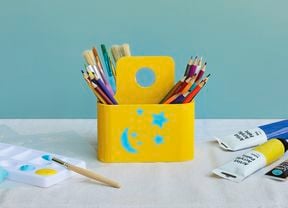 A bright yellow wooden art caddy decorated with blue stencilled stars and a moon, filled with coloured pencils and paintbrushes, surrounded by paint tubes and a paint palette.