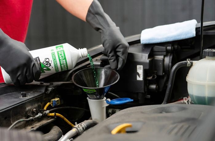 A person wearing gloves is pouring windshield washer fluid into a car's reservoir using a funnel.