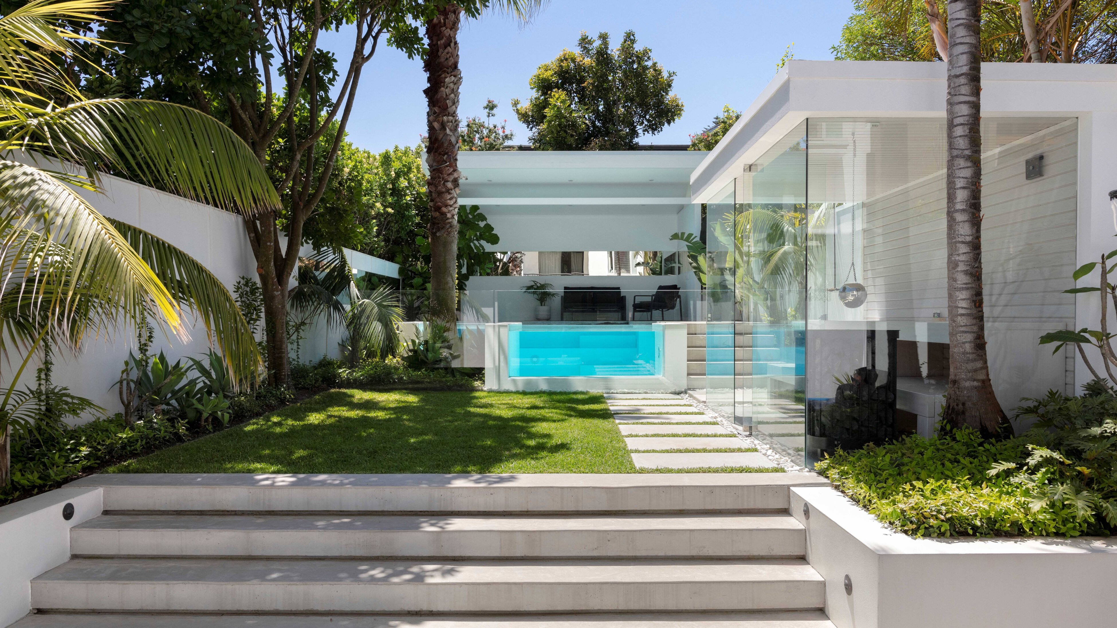 Modern backyard with wide concrete steps leading to a rectangular lawn, palm trees, a stone paving path with grass strips, and a glass‑walled pool