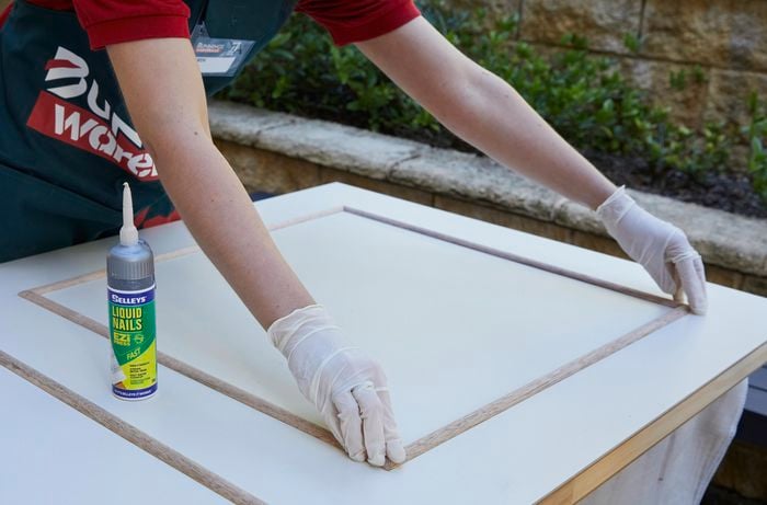 A person wearing disposable gloves uses Selleys Liquid nails construction adhesive to install moulding on a door