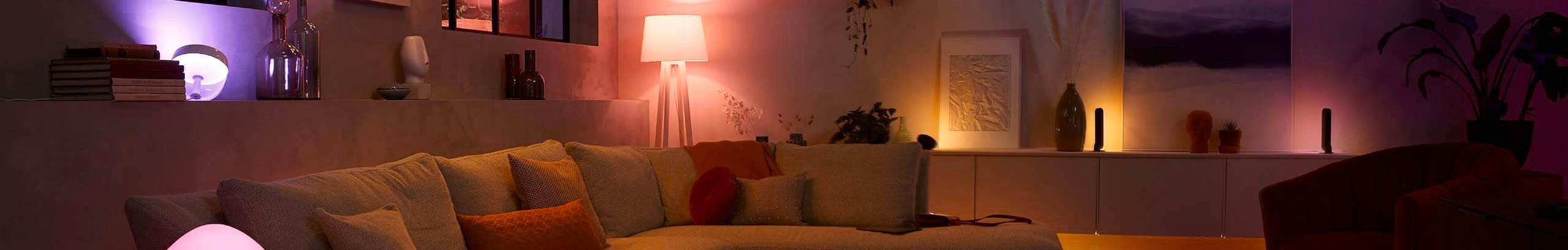 Modern living room illuminated with warm amber and purple smart lighting. A large sectional sofa with neutral and orange cushions sits in the foreground, surrounded by decorative shelves, table lamps, framed artwork, vases, and indoor plants. Soft ambient lighting creates a cozy, contemporary atmosphere throughout the space.