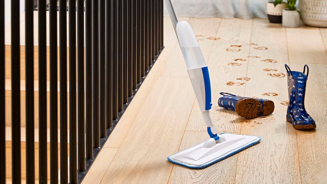 Oates Spray Mop leaning against stairwell railing, next to muddy gumboots on a timber floor.