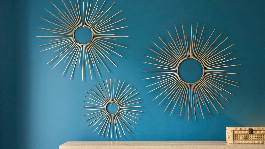 Three gold sunburst wall art pieces made from cable ties arranged on a teal wall, each featuring a circular center with evenly spaced radiating spokes in varying sizes, displayed above a light wooden surface with a woven storage box.