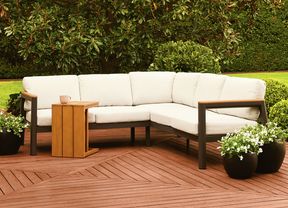 Outdoor corner lounge with a wooden side table on a timber deck, surrounded by green foliage.