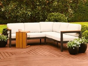 Outdoor corner lounge with a wooden side table on a timber deck, surrounded by green foliage.