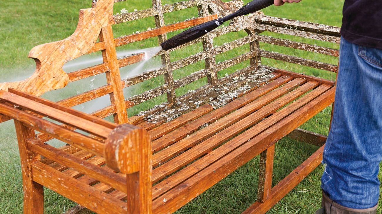 A person using a pressure washer to clean and restore a teak wood bench.