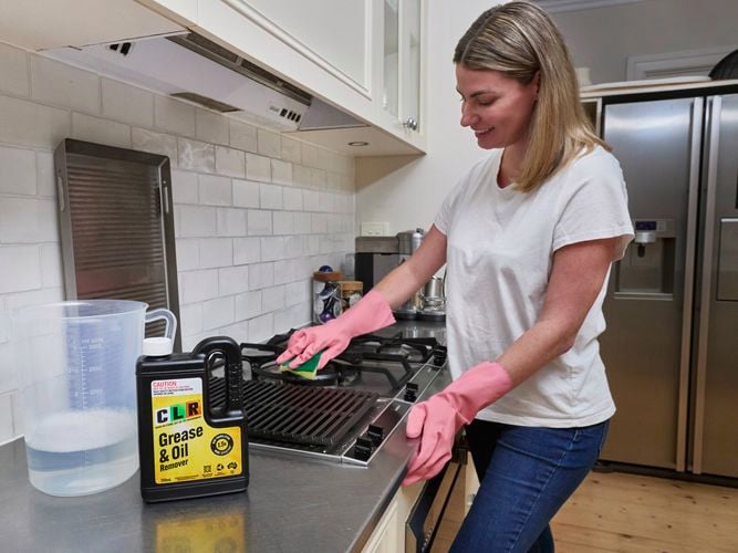 A lady in a kitchen holding a sponge against a stove top with a bottle of CLR Grease & Oil 750ml next to a jug of water.