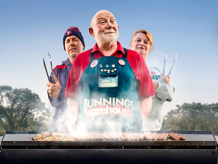 A Bunnings Team Member and 2 people in front of a BBQ with food on top of it.