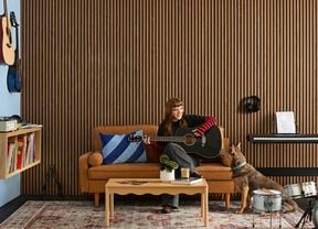 A music room with acoustic wall panelling, rubber floor matting, a tan sofa, drum kit, record player on floating shelves, wall-mounted guitars and a keyboard 