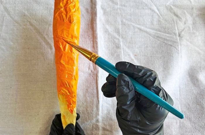 Black gloved hands hold and use a small paintbrush to paint a carrot made from masking tape in orange.