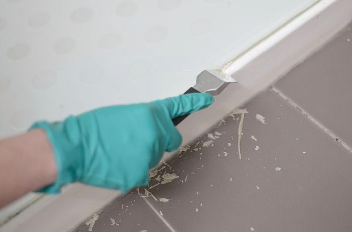 A hand wearing green gloves is using a scraper to remove silicone.