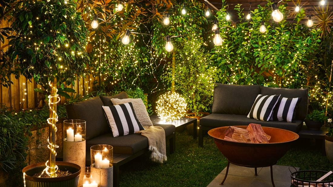A backyard lit by fairy lights and candles, with outdoor furniture surrounding a fire pit