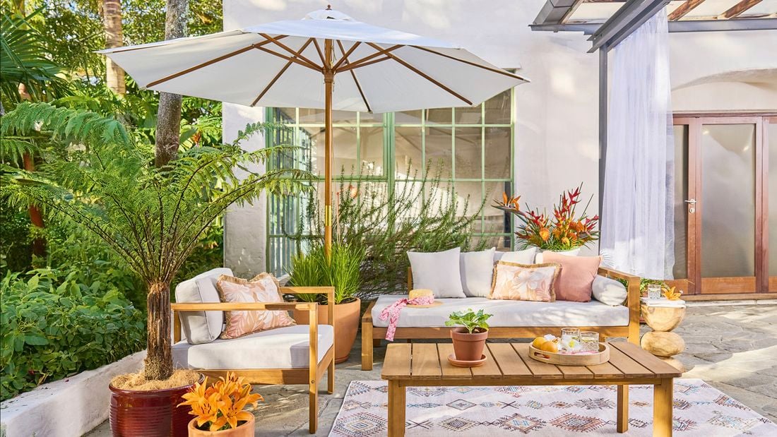 An outdoor patio setting with a raised bed to the left, a table, lounge with stylish cushions and a white and timber market umbrella.