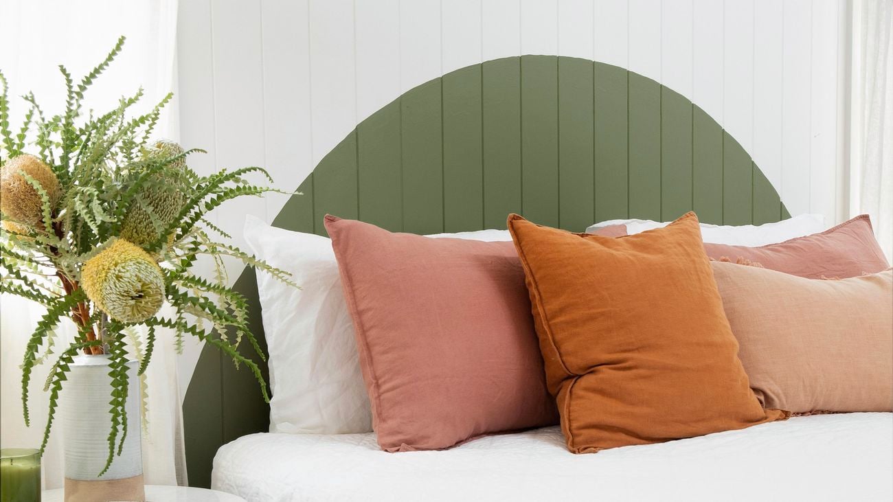 White bed with pink, white and orange pillows, a wooden bedside table, a pink throw blanket and a painted green headboard.