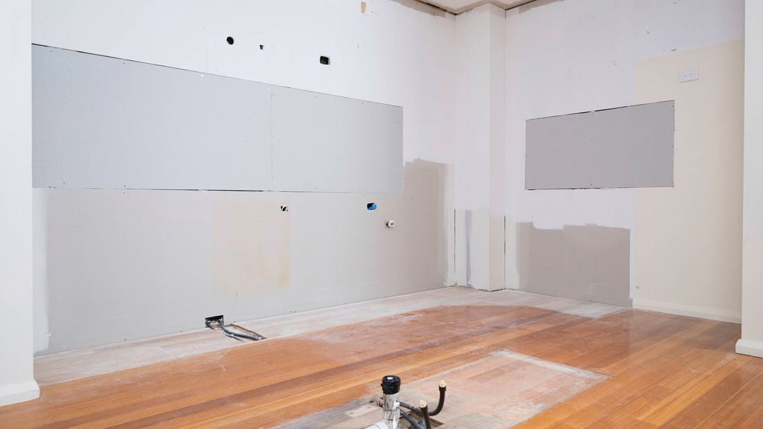 The image shows a completely stripped kitchen space with drywall patches and exposed flooring.