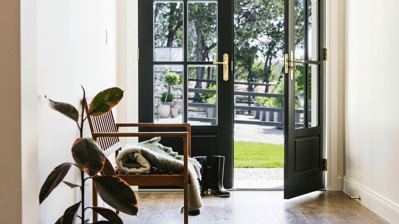 An entryway at the Little Lyndoch farmhouse with dark wooden floors, a wooden bench with a blanket, two potted plants, and open black French doors leading to an outdoor area