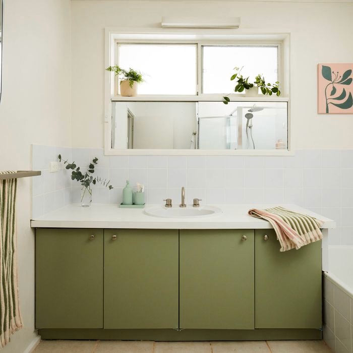 Bright bathroom with a green vanity cabinet and white benchtop, featuring a central sink beneath a wide mirror, a bathtub to the side, white tiled walls, striped towel on a rack, and potted plants along the windowsill for natural light and greenery.