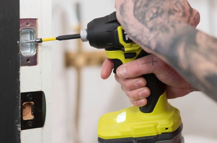 A person is using a drill to secure a strike plate on a door frame