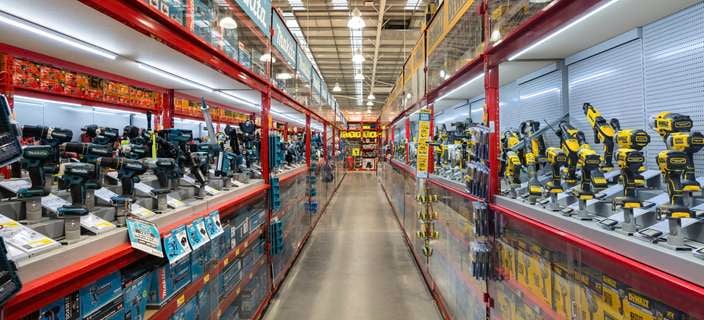 Wide aisle inside a hardware store displaying rows of power tools; Makita cordless drills and equipment in teal on the left and DeWalt tools in yellow and black on the right, arranged on lit shelves with boxed stock below and signage above.