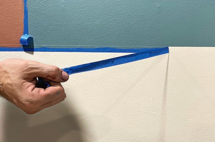 Close up of a hand removing blue painter’s tape from a freshly painted wall that shows three colours