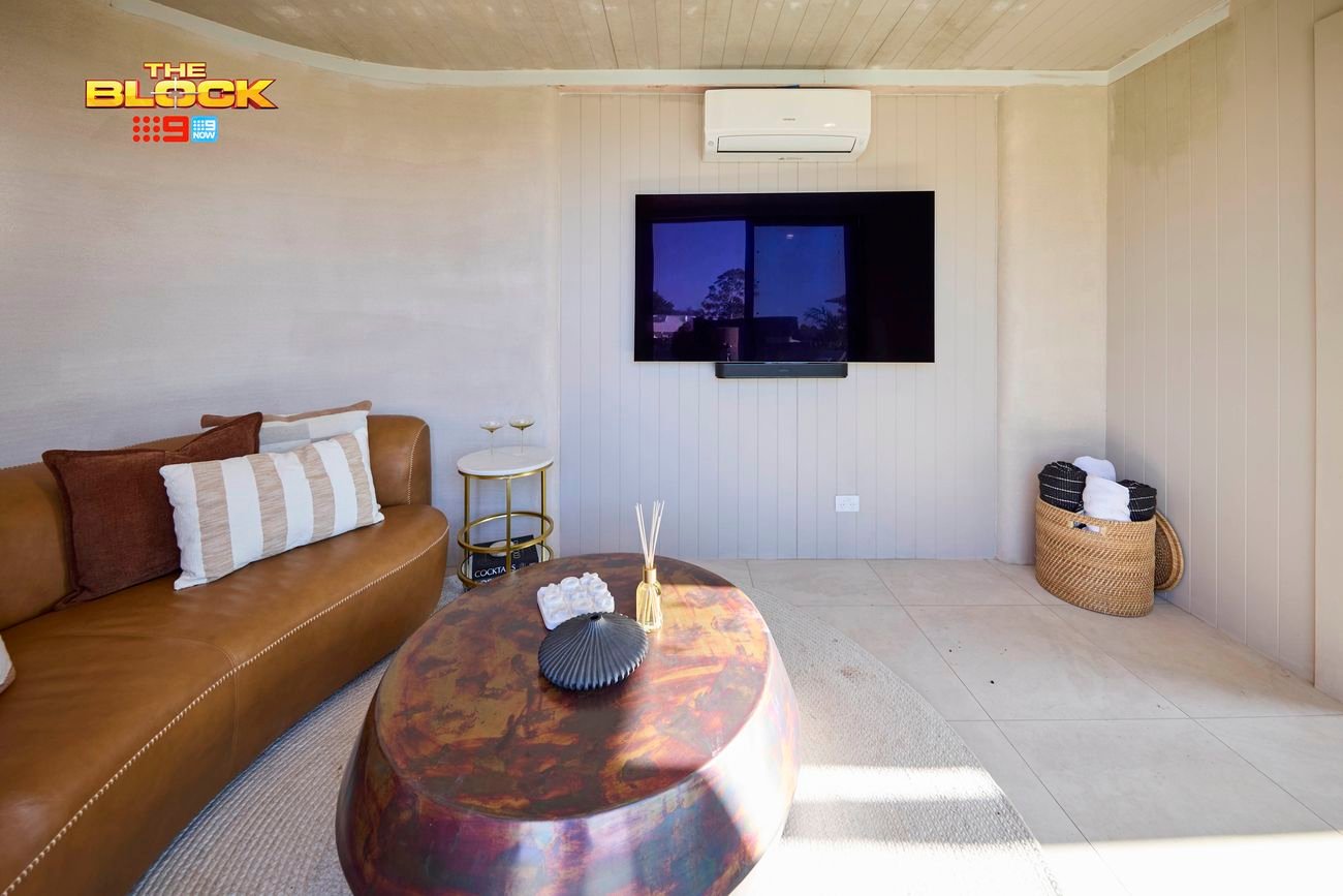 A room with a couch with cushions on it, a table, a tv mounted to a wall and a stool in it.