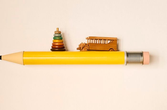 A large yellow pencil shelf with a pink eraser end mounted on a neutral wall, with wooden toys on top.