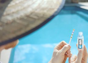 Person holding a swimming pool water testing strip and test solution bottle beside a blue swimming pool.
