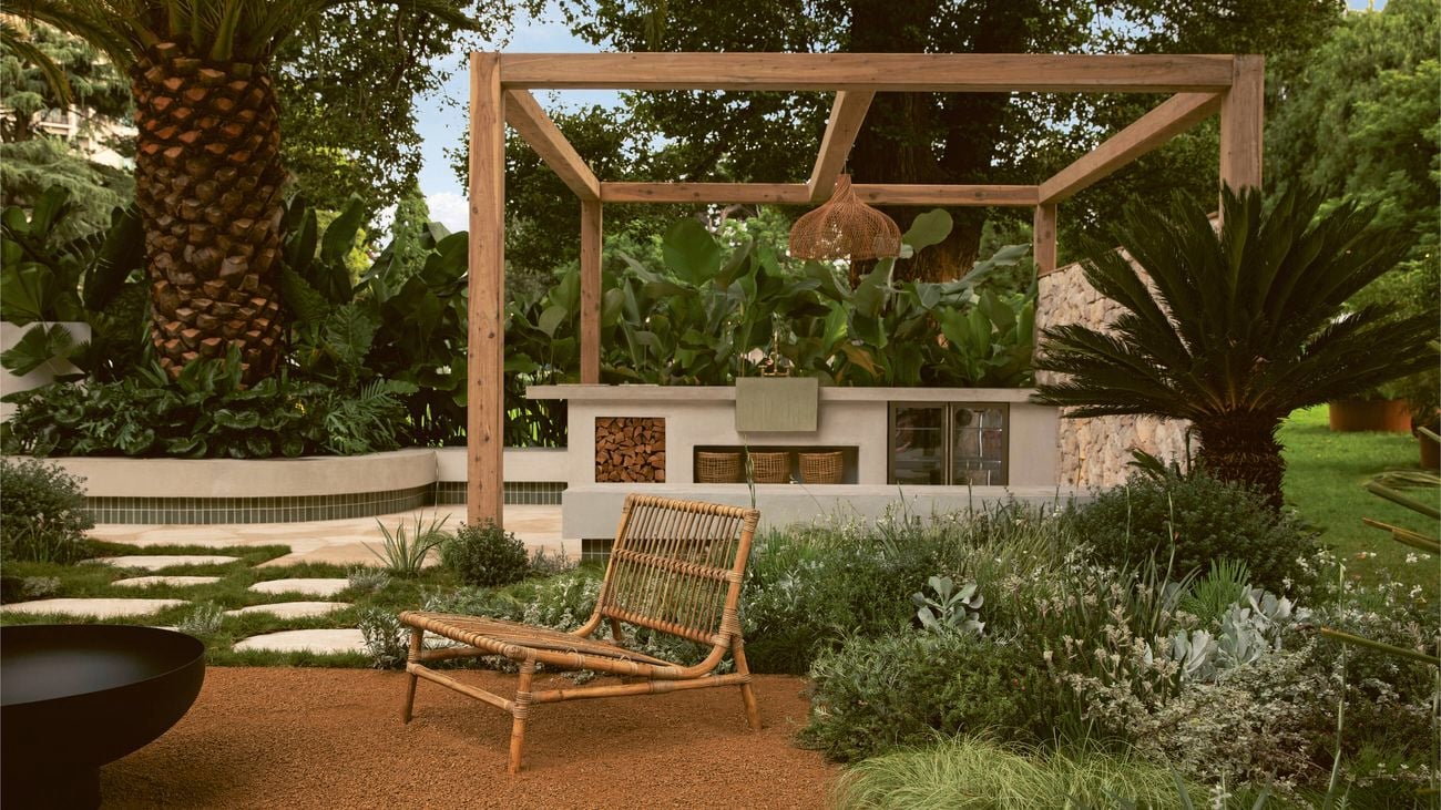 Woven lounge chair in a landscaped garden with a timber pergola and outdoor kitchen.