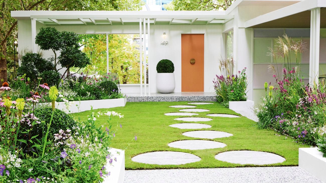  A front garden of a house with a round stepping stone path in the lawn leading to a front door, with lushly planted garden beds around the edges and to the right a panelled garage door painted in pastel shades.