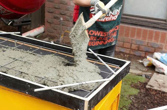 Person using shovel to scoop concrete into form ply frame with reinforcing mesh.