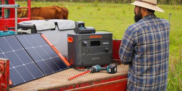 : The back of a male wearing a wide-brimmed hat and a check shirt standing in front of a Jackery portable power station on the back of a ute in a farm setting.