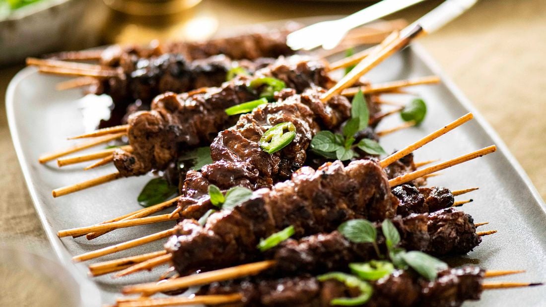 A white platter piled with BBQ skewers garnished with sliced green chilli and oregano leaves.