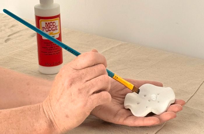 Hands applying sealant with a paintbrush to a circular piece of clay with holes, with a bottle of sealer in the background.