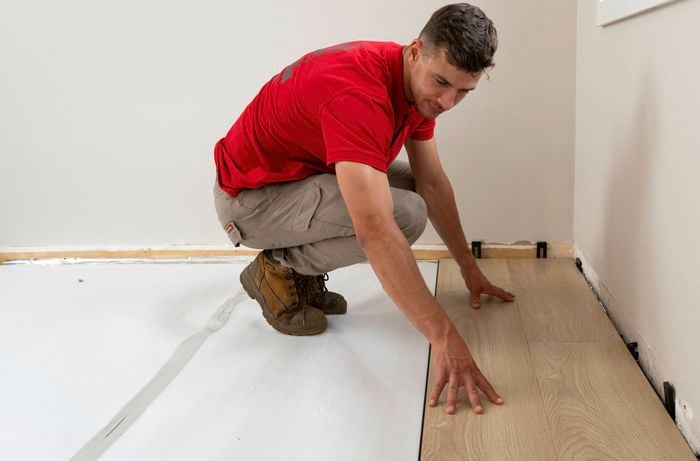 A Bunnings Team Member is carefully installing laminate flooring positioning a plank near the wall