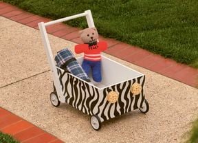 : A zebra-print toy wagon with a teddy bear and a plaid blanket on a paved path next to a lawn.