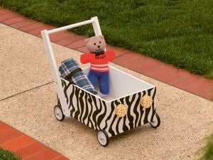 : A zebra-print toy wagon with a teddy bear and a plaid blanket on a paved path next to a lawn.
