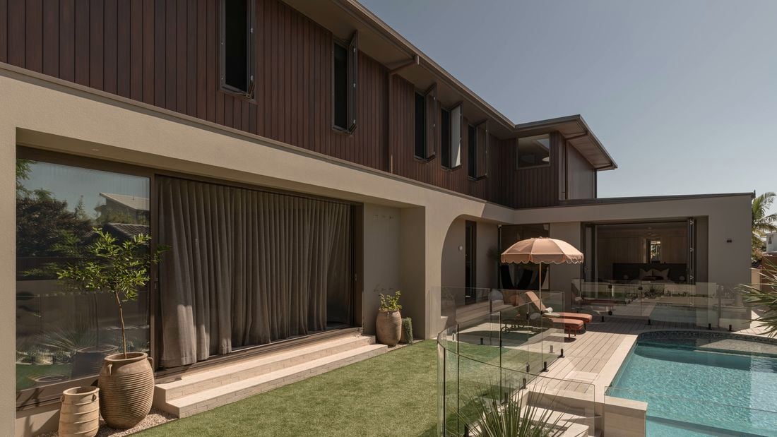 Contemporary two-storey house with timber cladding and rendered walls overlooking a backyard with a glass-fenced swimming pool, patio seating, and landscaped garden.