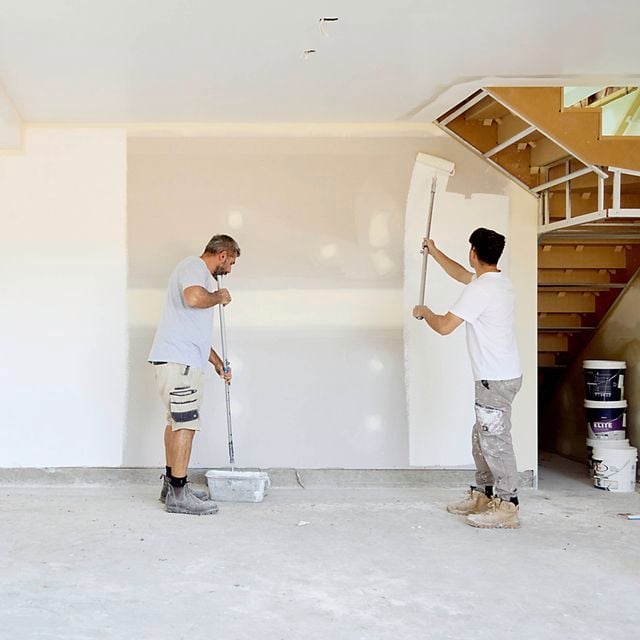 Two people painting interior walls of an unfinished room, using long-handled rollers. The space has bare concrete flooring, partially painted white walls, exposed sections around windows, and a wooden staircase with construction materials and paint buckets nearby.