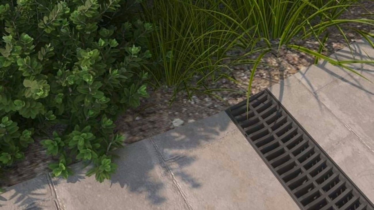 Concrete pavers with a linear drainage grate beside garden plants and gravel edging.