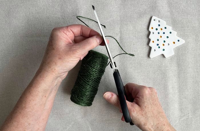 A pair of hands cutting green twine with scissors next to a clay Christmas tree ornament on a drop sheet.