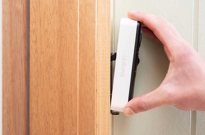 A person is attaching a Swann security doorbell onto the mounted bracket on a wooden door frame