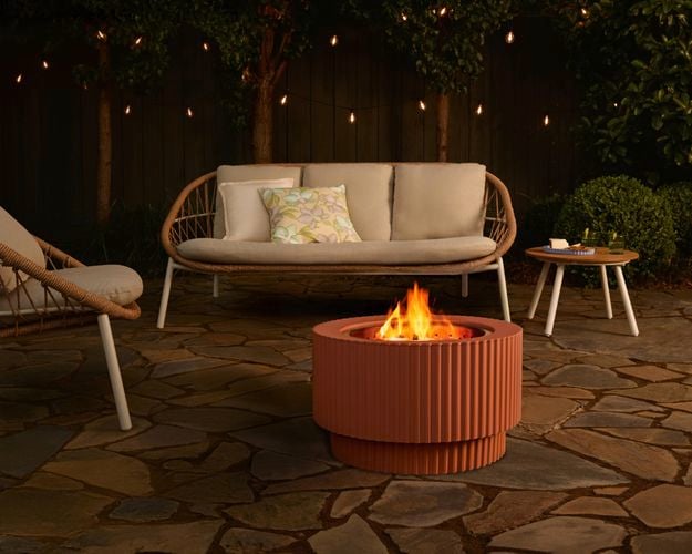 Round terracotta outdoor fire pit with a visible flame, set on a stone patio and surrounded by outdoor lounge seating, string lights and garden trees at night.