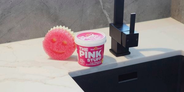 A pink Scrub Mommy sponge and a tub of The Pink Stuff cleaning paste sitting on a benchtop beside a black tap.