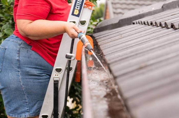 A person in a red shirt is standing on a ladder, using a garden hose to flush out a roof gutter after cleaning.