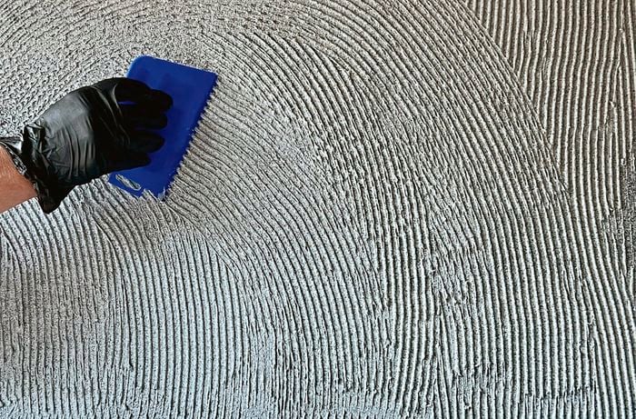 A person wearing a disposable glove uses a notched trowel to spread tile adhesive over a wall.