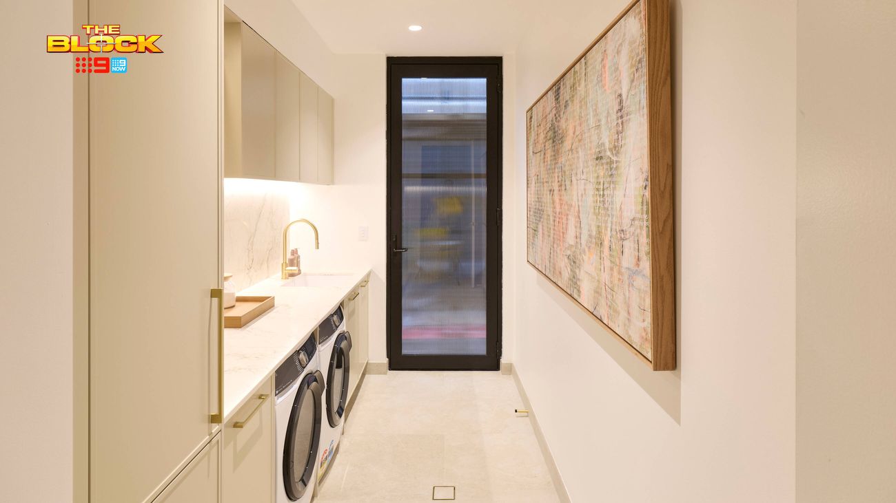 Modern laundry featuring built-in cabinetry, stone benchtop, front-loading appliances, gold tapware and framed artwork.