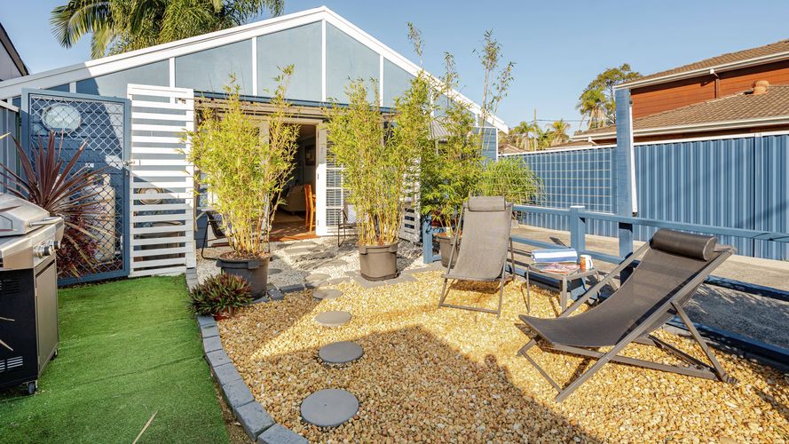 Private outdoor courtyard with gravel landscaping, potted bamboo, sun loungers and a barbecue, leading to a light blue beach-style cottage with open doors.