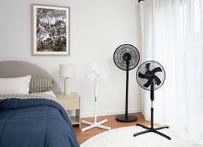 Three pedestal fans in white and black positioned beside a bed in a bright bedroom with sheer curtains and a bedside table.